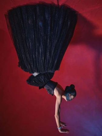A woman in a black tulle dress performs an acrobatic handstand against a red backdrop with dramatic lighting, showcasing a blend of fashion and fitness.
