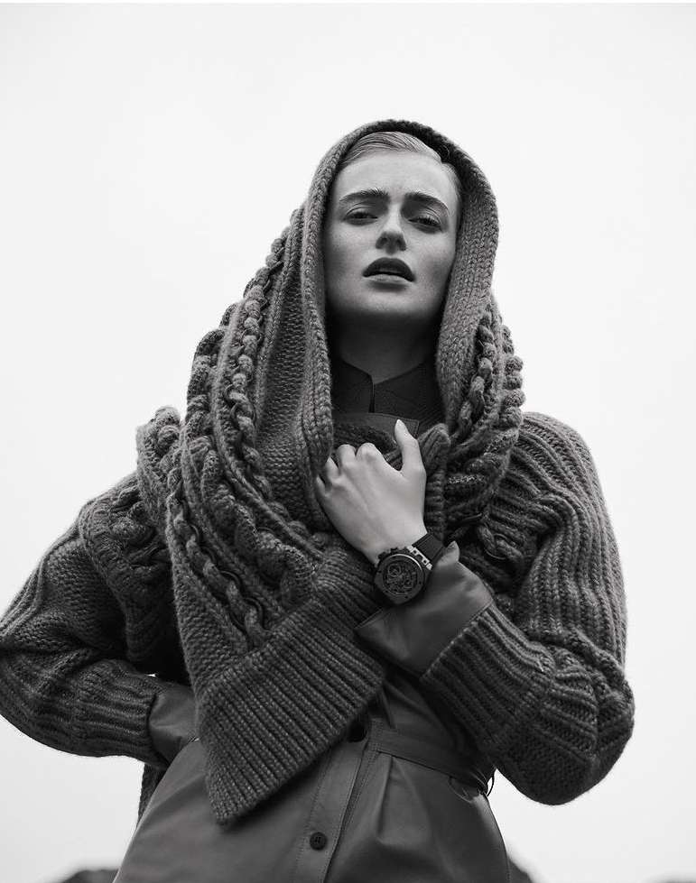 Monochrome portrait of a woman with a neutral expression wearing a knitted hood and a leather-like coat against a light background.