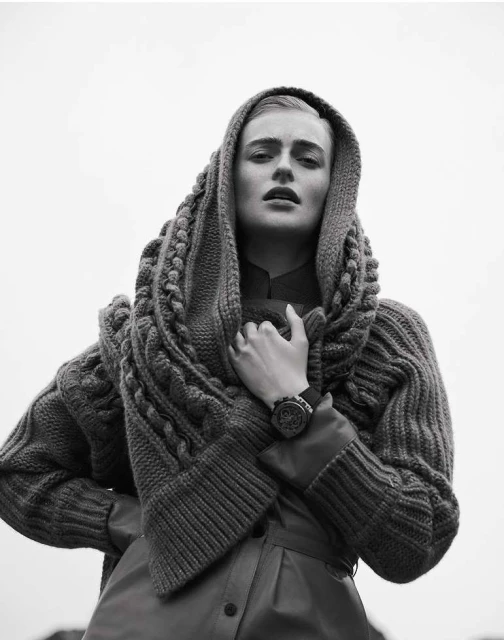 Monochrome portrait of a woman with a neutral expression wearing a knitted hood and a leather-like coat against a light background.