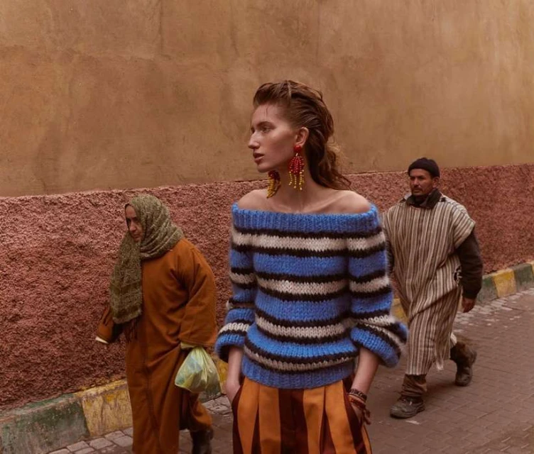 Fashion editorial shot of a woman in a striped sweater, orange skirt, and beaded earrings, walking in an alleyway with other pedestrians.