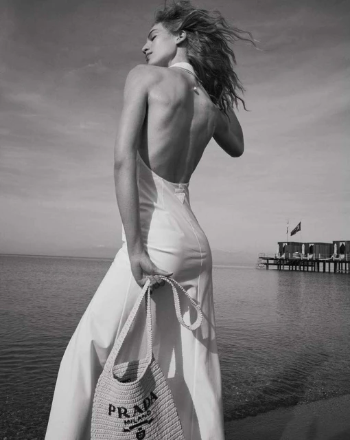 Black and white portrait of an adult woman on a beach, wearing a dress and holding a Prada handbag with a pier visible in the background.