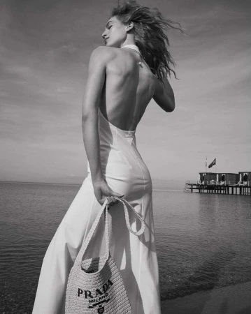 Black and white portrait of an adult woman on a beach, wearing a dress and holding a Prada handbag with a pier visible in the background.