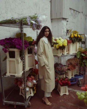 A brunette woman in a beige coat stands amid a display of colorful flowers, with a concrete wall and a metal shelving unit in the background.
