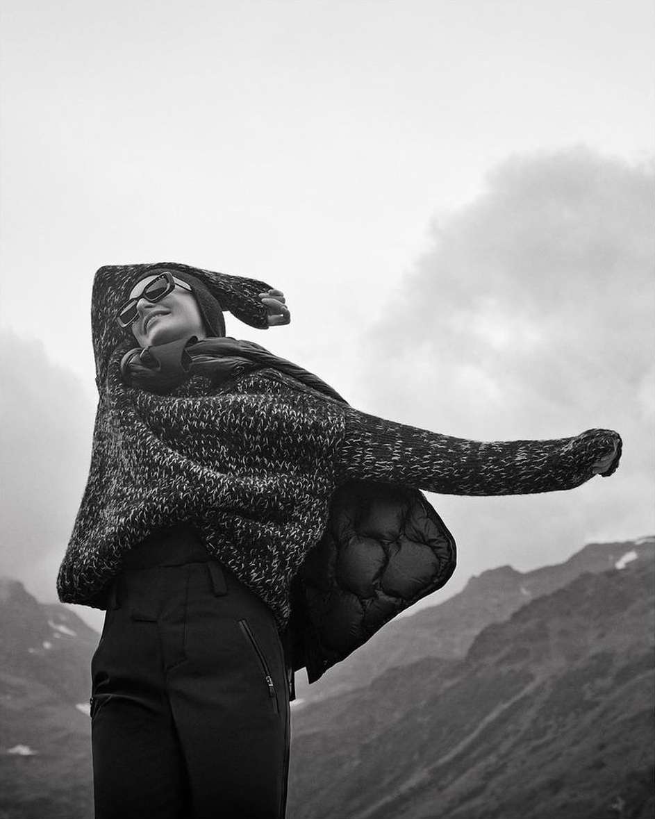 Black and white portrait of a woman wearing sunglasses and a textured sweater with arms raised against mountains