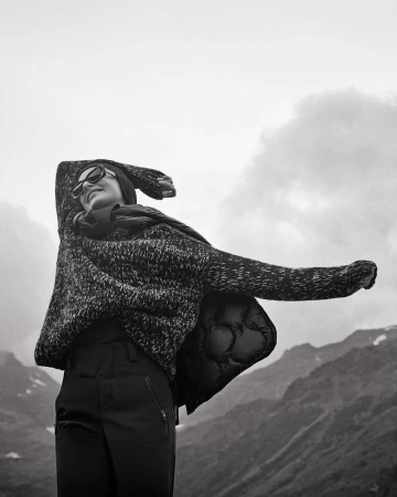 Black and white portrait of a woman wearing sunglasses and a textured sweater with arms raised against mountains
