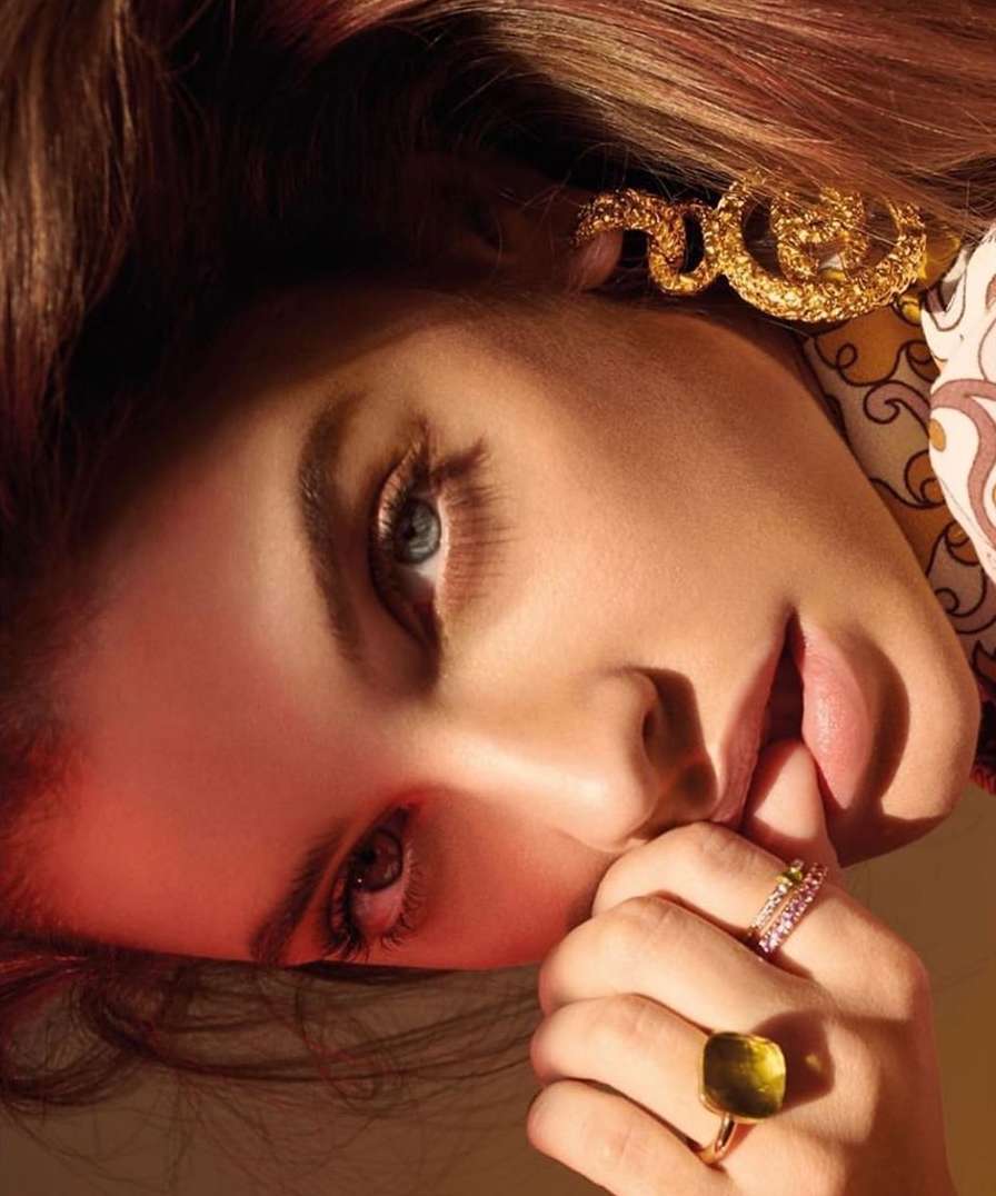 Close-up portrait of a light-skinned female model with light eyes, wearing gold jewelry and rings, holding fingers to her face.