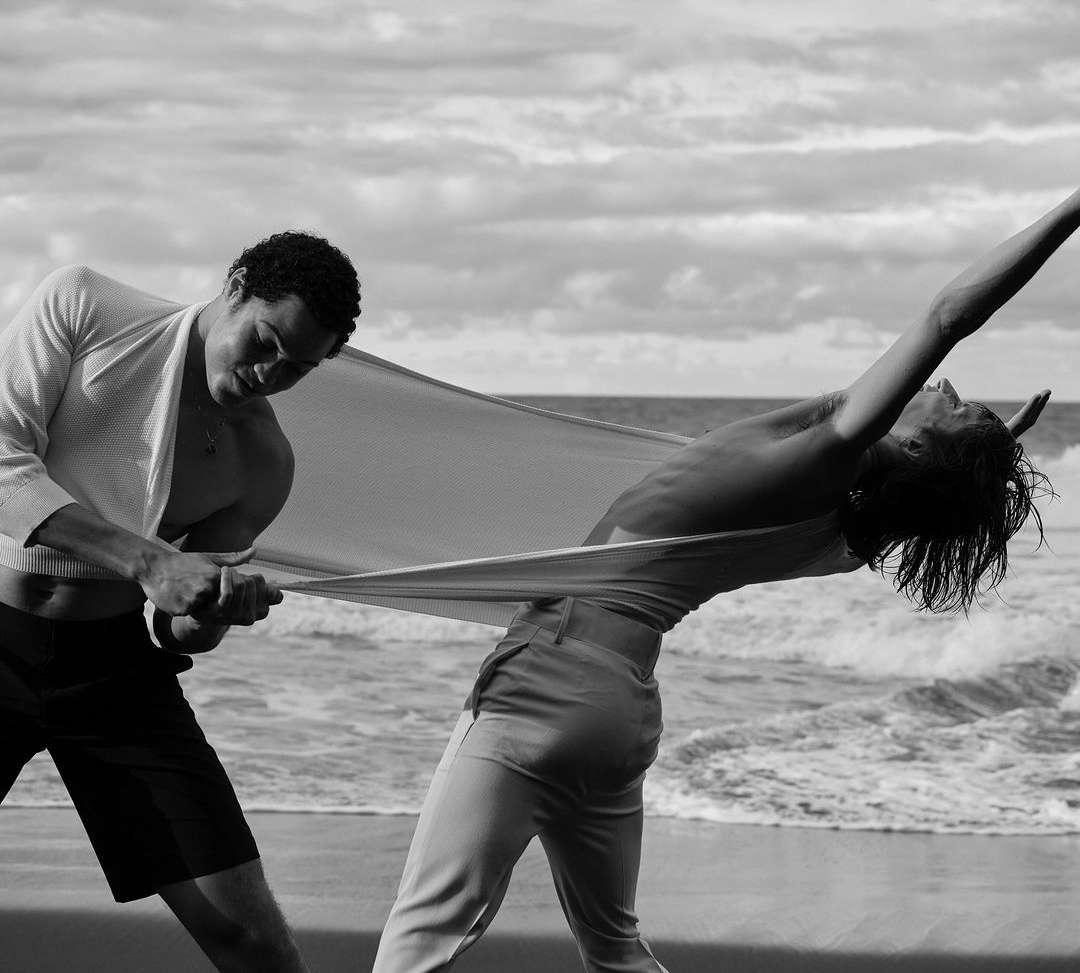 Two athletic people hold a sheet on a beach with an ocean and cloudy sky in the background, shot in black and white for fashion.
