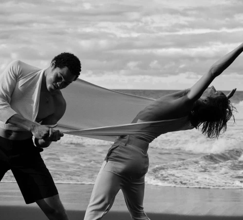 Two athletic people hold a sheet on a beach with an ocean and cloudy sky in the background, shot in black and white for fashion.