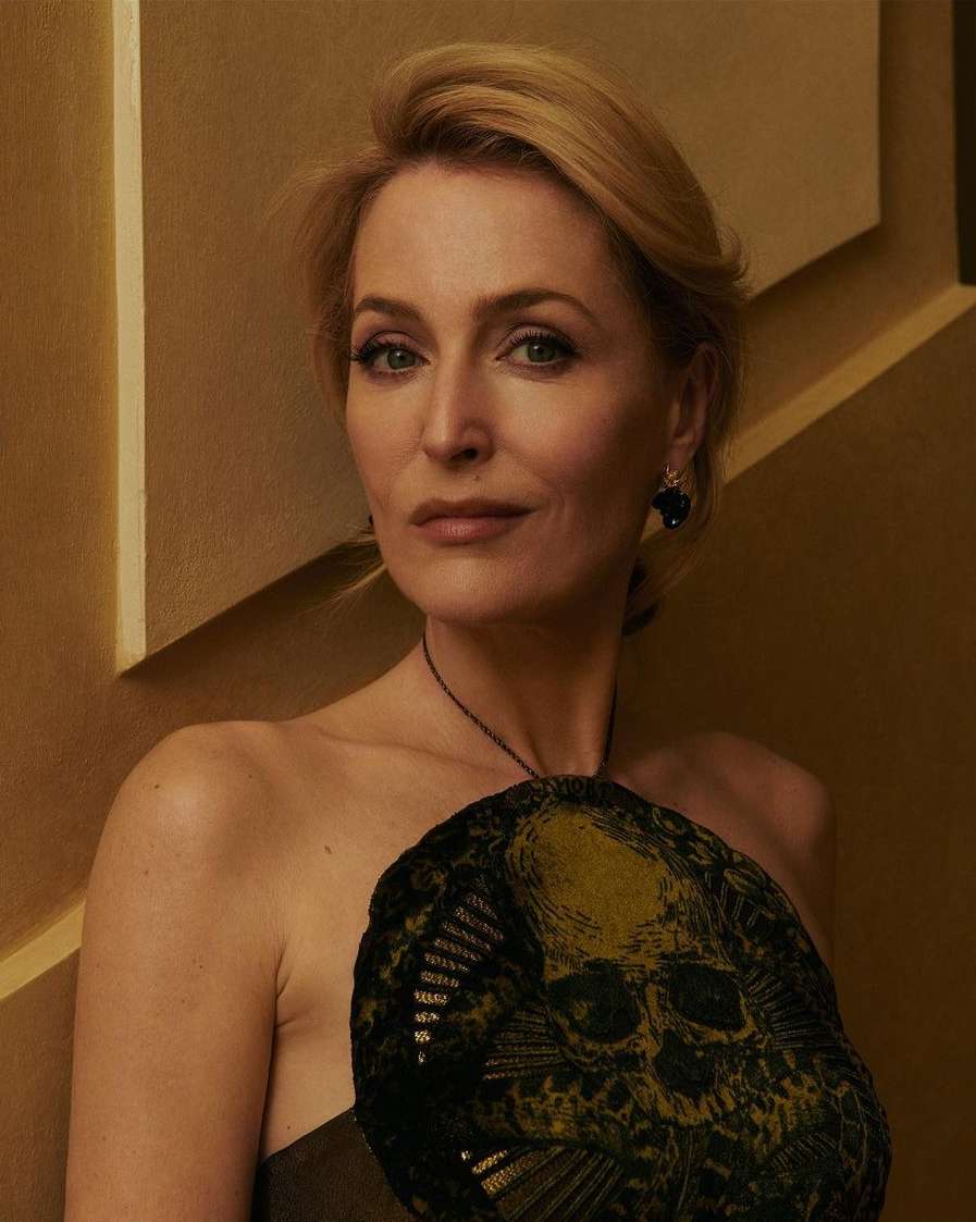 Close-up portrait of a blonde woman with shoulder-length hair in front of a tan background, wearing a strapless top and skull necklace.