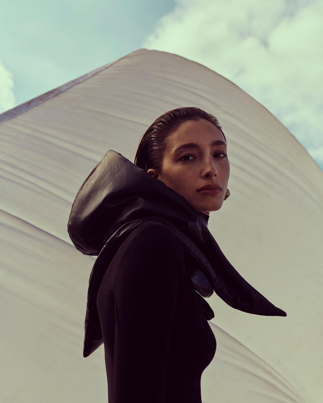 Fashion shot of a woman in a black outfit and hood posing against a white fabric and sky background, captured in a medium shot.