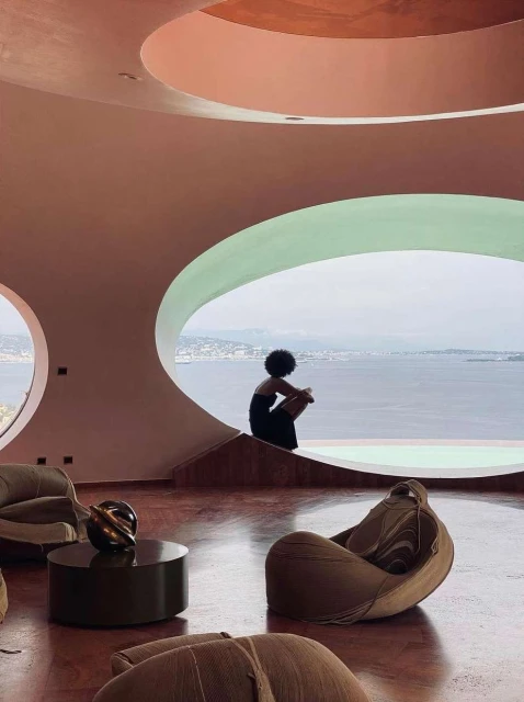 A woman in a black dress sits in a modern living room with large, round windows overlooking the ocean. The interior features a round table.