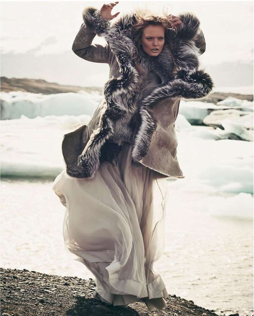 Woman in fur-trimmed coat and gown poses on rocky beach with glacial icebergs and cloudy sky in an outdoor fashion editorial shot.