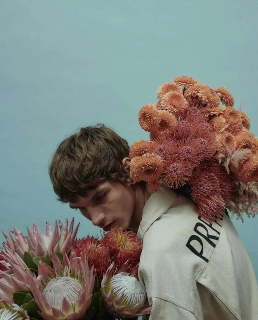 Young adult poses indoors among assorted protea flowers in a fashion shot with a teal background and light tan hoodie.