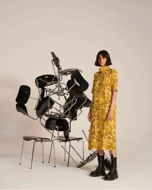 A woman in a yellow floral dress and black boots stands next to a precarious stack of black chairs in a minimalist studio setting.