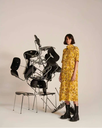A woman in a yellow floral dress and black boots stands next to a precarious stack of black chairs in a minimalist studio setting.