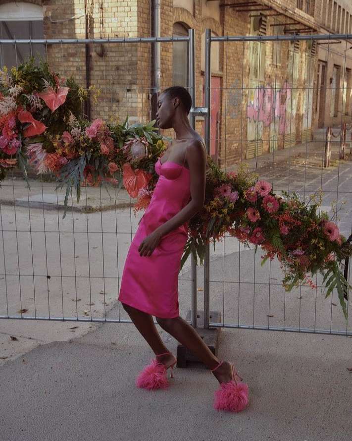 A dark-skinned woman in a pink dress and pink fuzzy heels poses next to a flower-covered construction fence in an urban setting.