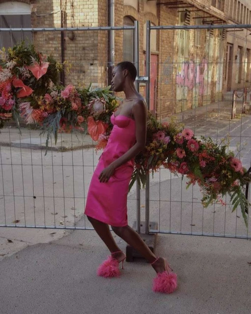A dark-skinned woman in a pink dress and pink fuzzy heels poses next to a flower-covered construction fence in an urban setting.