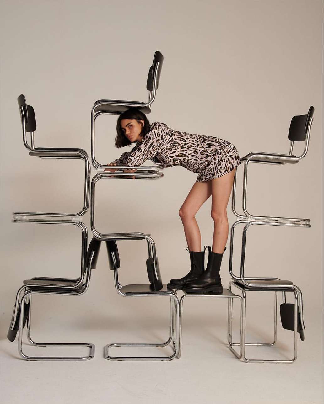 Fashionable young woman in a leopard print dress and black boots poses creatively on a stacked formation of chrome and black chairs in studio setting.