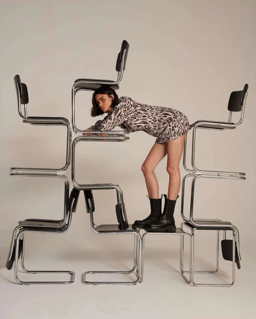 Fashionable young woman in a leopard print dress and black boots poses creatively on a stacked formation of chrome and black chairs in studio setting.