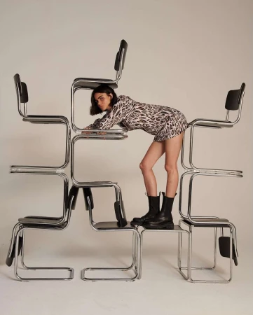 Fashionable young woman in a leopard print dress and black boots poses creatively on a stacked formation of chrome and black chairs in studio setting.