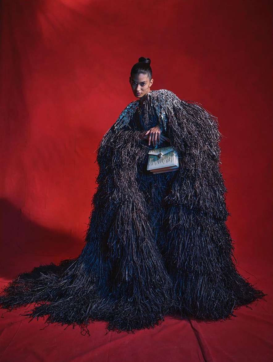 Fashionable woman in a black feathered cloak poses against a red backdrop, holding a boxy handbag with intricate gold-tone detailing.