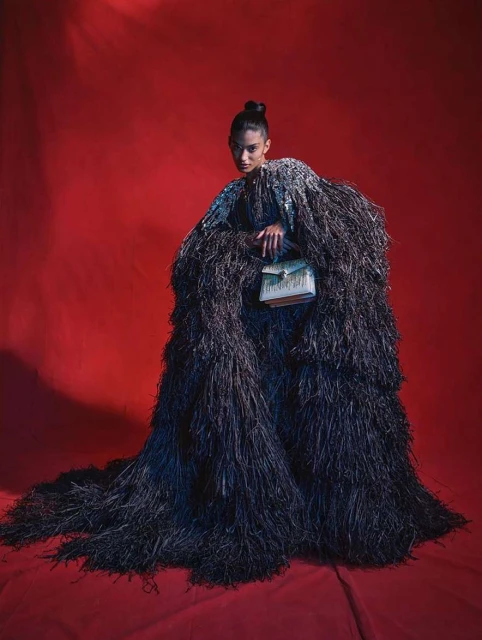 Fashionable woman in a black feathered cloak poses against a red backdrop, holding a boxy handbag with intricate gold-tone detailing.