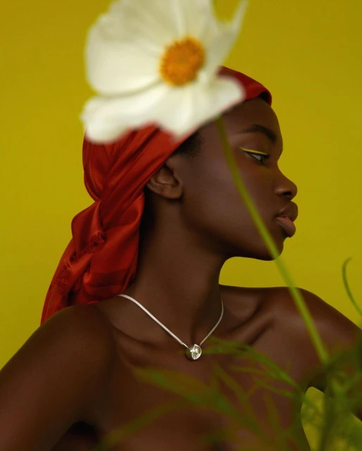 Portrait of a young Black woman in a headscarf and necklace, posing against a yellow backdrop with a white flower detail.