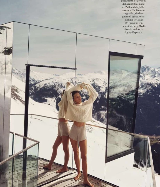 A woman in loungewear stands on a balcony against a snowy mountain backdrop, mirrored reflection echoing the alpine scenery.