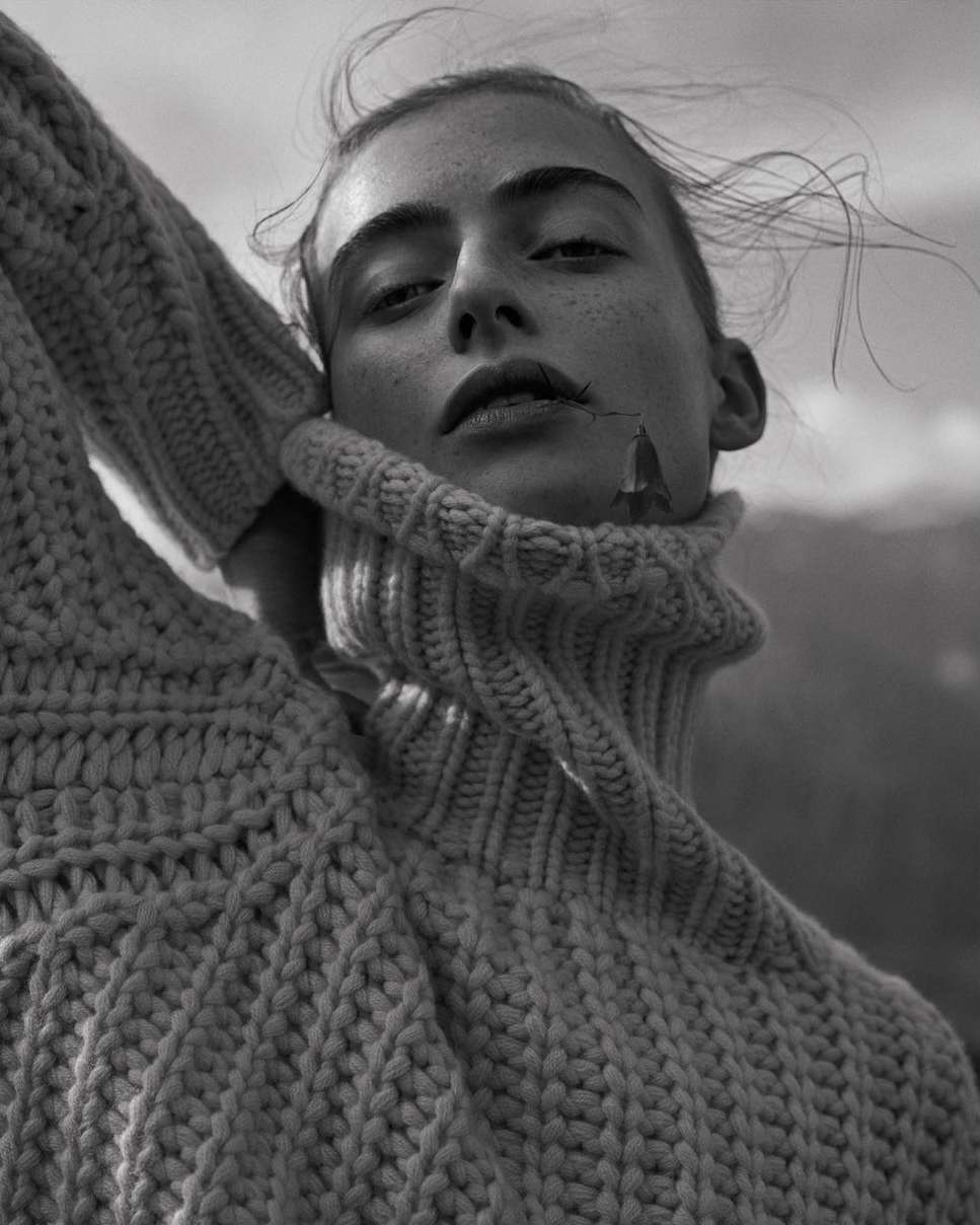 Monochrome portrait of a young woman with wind-blown hair, wearing a chunky knit turtleneck sweater, against a blurred mountain backdrop.