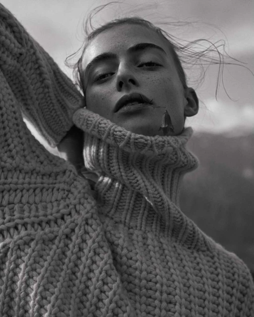 Monochrome portrait of a young woman with wind-blown hair, wearing a chunky knit turtleneck sweater, against a blurred mountain backdrop.
