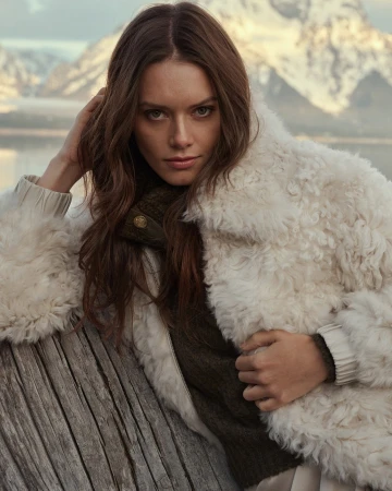 Woman wearing a textured white faux fur jacket leaning against a rustic wooden post with a snowy mountain background.