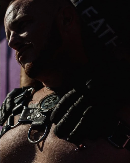 Close-up of a bald, muscular man with a mustache wearing black leather harness with metal hardware and black leather gloves in a dimly lit indoor space.