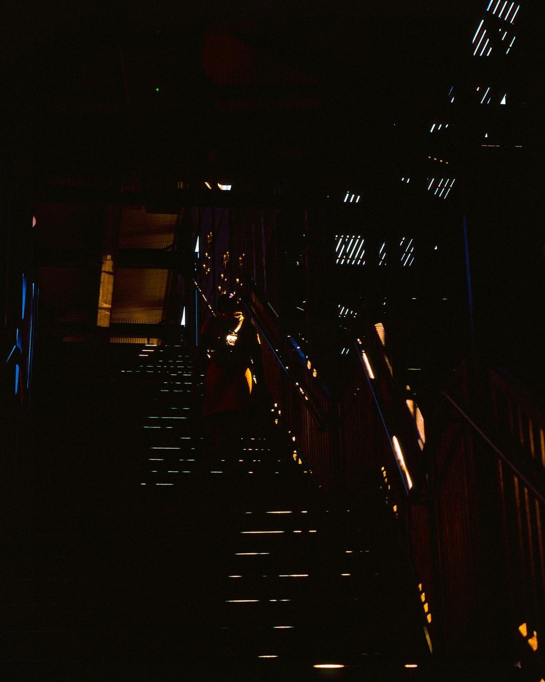 Dimly lit staircase with handrails, illuminated by scattered lights in an indoor space, creating a stark contrast with dark surroundings.