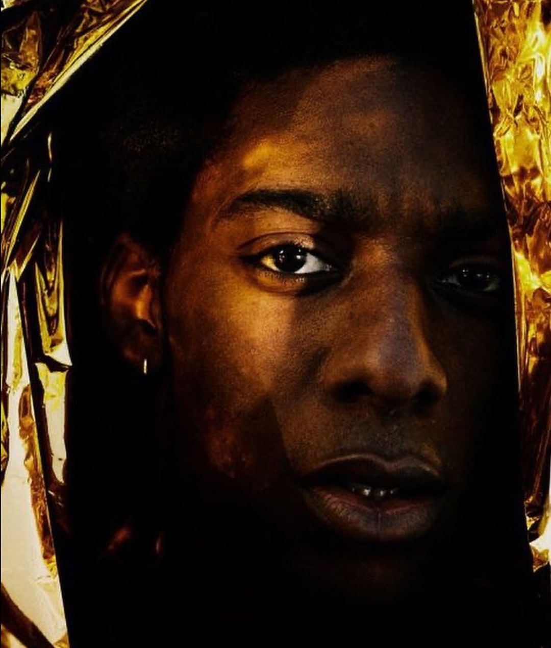 Close-up portrait of an adult male with dark skin, partially obscured by shimmering gold fabric, creating dramatic light and shadows.