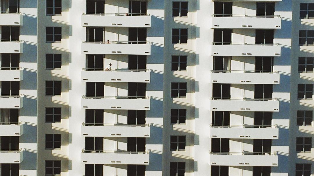 Exterior of high-rise building with rows of balconies and windows, featuring two figures visible on separate balconies in daylight.