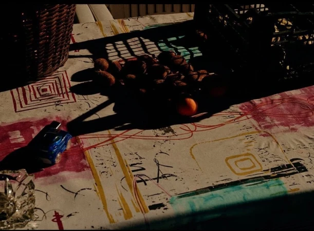 Still life on a patterned tablecloth features walnuts, orange, and basket under shadows, with a toy car and glass dish in the foreground.