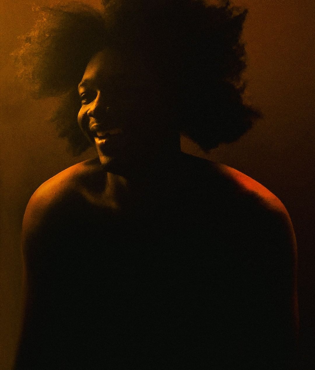 Portrait of an African American adult male with an afro, smiling in a close-up shot with dramatic yellow lighting and a dark background.