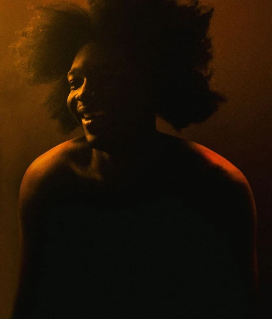 Portrait of an African American adult male with an afro, smiling in a close-up shot with dramatic yellow lighting and a dark background.
