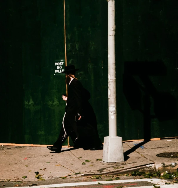 A man in traditional Hasidic Jewish clothing walks along a city sidewalk next to a grey utility pole and a green boarded-up wall.