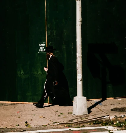 A man in traditional Hasidic Jewish clothing walks along a city sidewalk next to a grey utility pole and a green boarded-up wall.