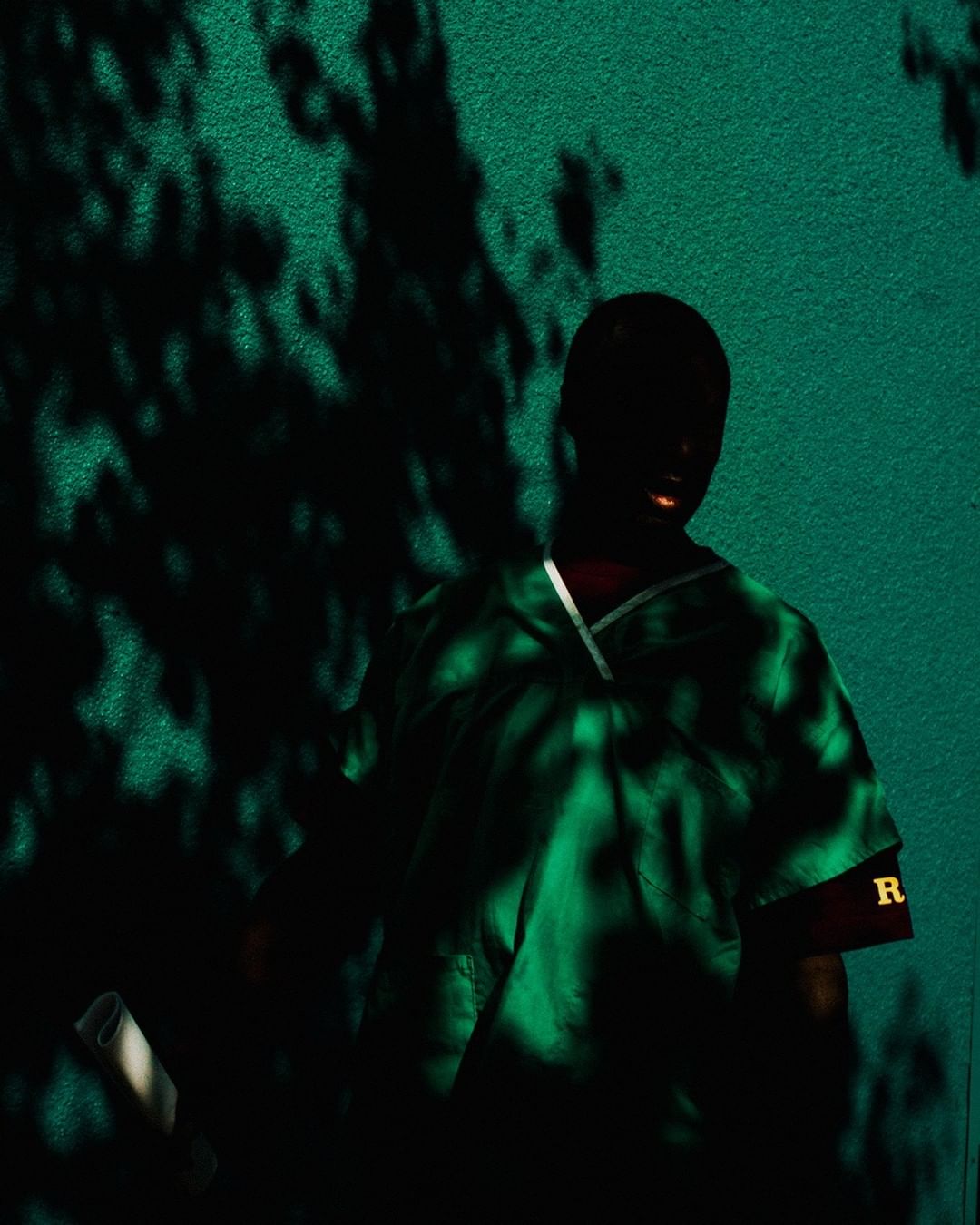 A person stands in a green scrub suit against a green wall, with shadow patterns from trees or foliage, with the letter R on the sleeve.