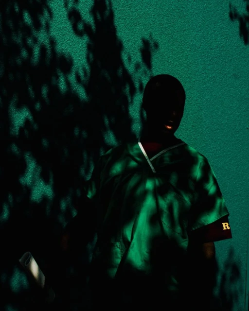 A person stands in a green scrub suit against a green wall, with shadow patterns from trees or foliage, with the letter R on the sleeve.