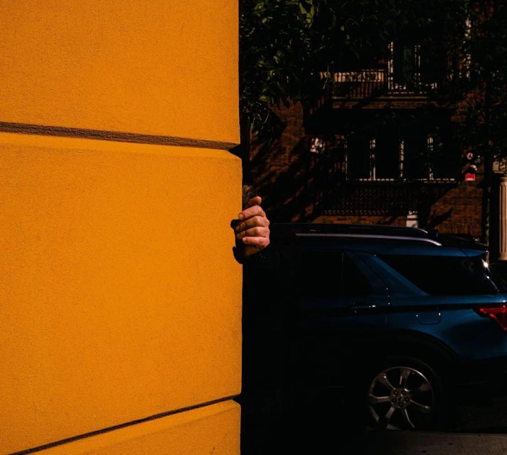 Hand peeks from behind a bright yellow building wall. A dark blue SUV is parked nearby in an urban setting with leafy trees.