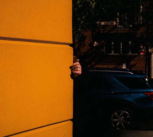 Hand peeks from behind a bright yellow building wall. A dark blue SUV is parked nearby in an urban setting with leafy trees.
