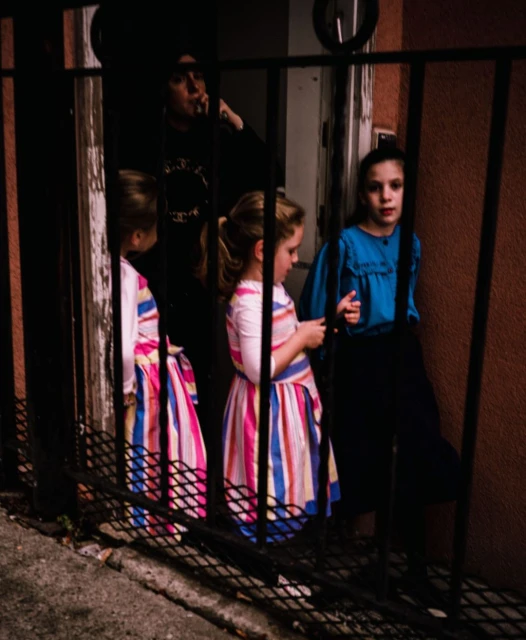 Low-angle shot of a group of children standing behind a black metal fence on a city street with an adult in the background.