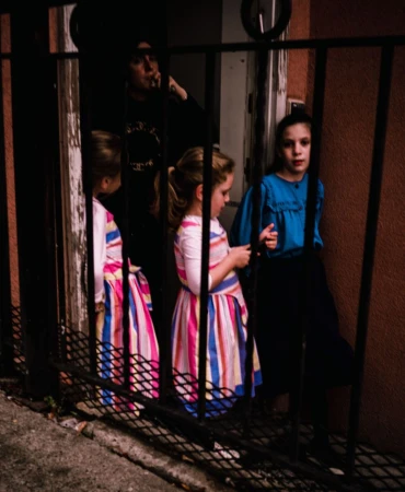 Low-angle shot of a group of children standing behind a black metal fence on a city street with an adult in the background.