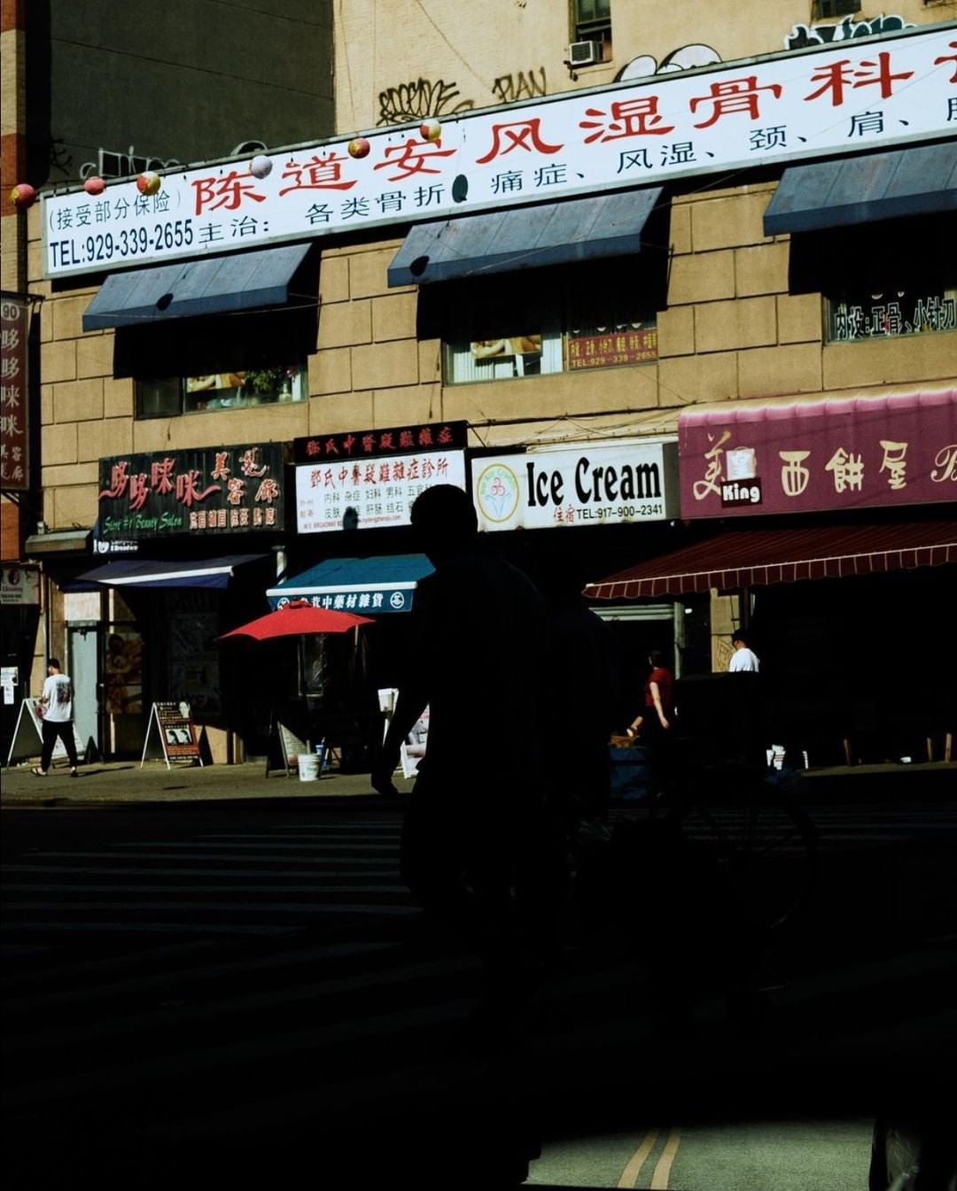 Street scene featuring a silhouetted figure crossing a crosswalk in front of Asian-style businesses in a brightly lit urban environment.