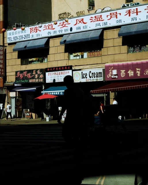 Street scene featuring a silhouetted figure crossing a crosswalk in front of Asian-style businesses in a brightly lit urban environment.
