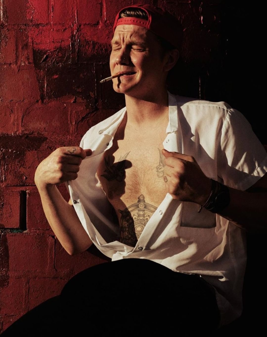 Man with tattoos poses against red brick wall in white shirt and red hat, smoking and looking up, with chest tattoos visible.