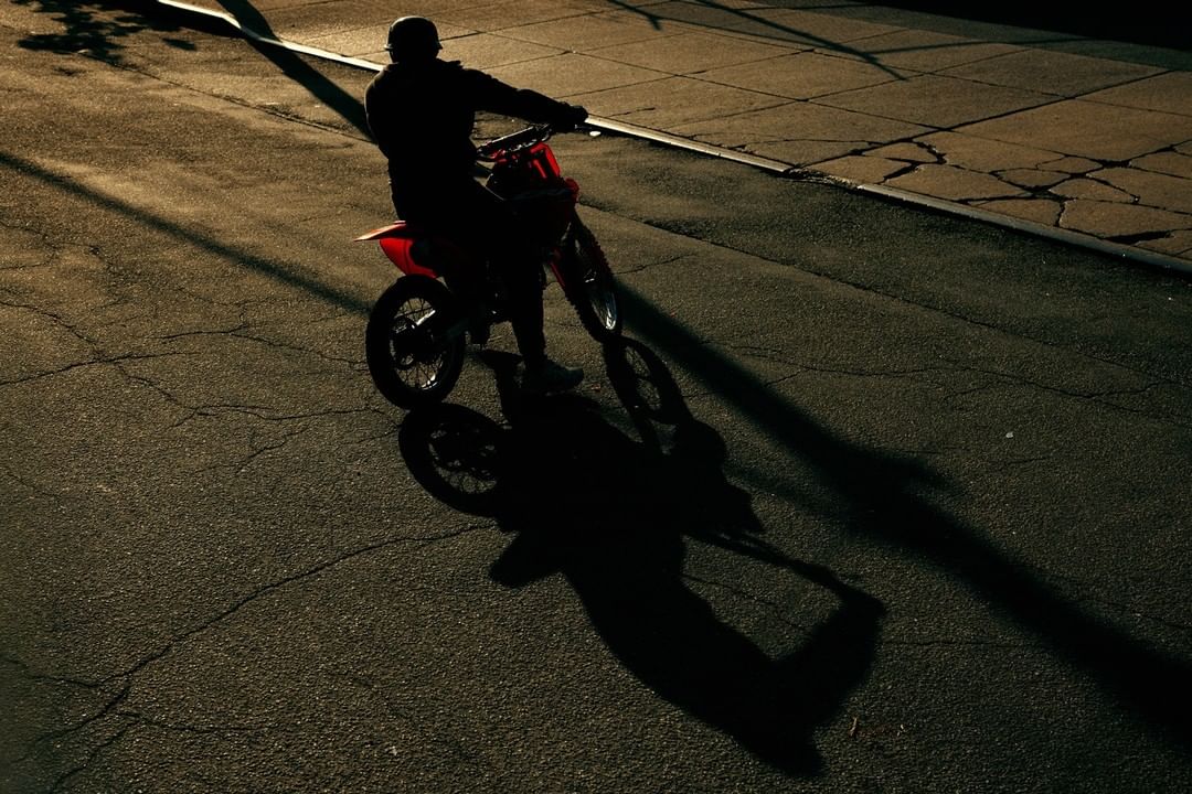 A person rides a red dirt bike on an asphalt road at dusk, creating a silhouette and long shadow. The scene is moody and urban.